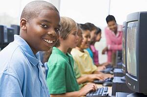 Six children at computer terminals with teacher in background (depth of field/high key)