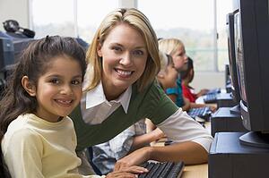 Teacher helping student at computer terminal with students in background (depth of field/high key)