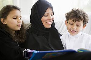 Mother and two young children in living room reading book and smiling (high key/selective focus)