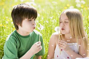 Two young children sitting outdoors blowing dandelion heads smiling