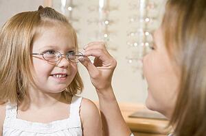 Woman trying eyeglasses on young girl at optometrists smiling