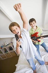 Two Boys Standing On A Bed, Playing Guitar And Singing Into A Hairbrush