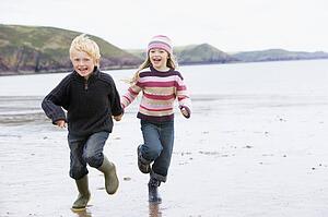Two young children running on beach holding hands smiling