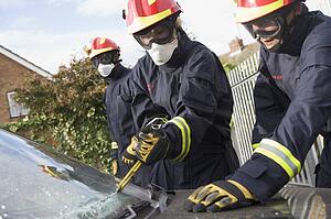 Two firefighters cutting out a windshield after an accident with another firefighter in background