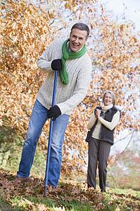 Man outdoors raking leaves and woman in background smiling (selective focus)
