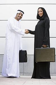 Two businesspeople standing outdoors with briefcases shaking hands and smiling