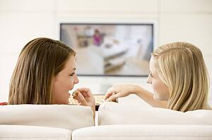 Two women in living room watching television eating chocolates smiling