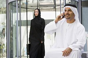 Businessman sitting outdoors by building using cellular phone with businesswoman walking in background (selective focus)