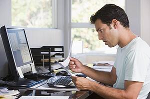 Man in home office using computer holding paperwork and looking shocked