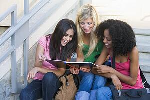 Three women sitting on staircase outdoors looking at notebook (high key)