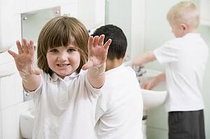 Students in bathroom at sinks washing hands with one holding up soapy hands (selective focus)