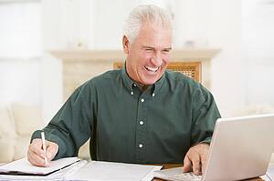 Man in dining room with laptop and paperwork smiling