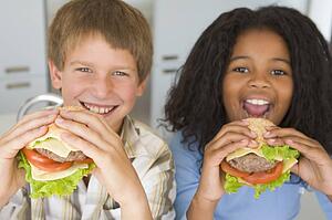 Two young children in kitchen eating cheeseburgers smiling