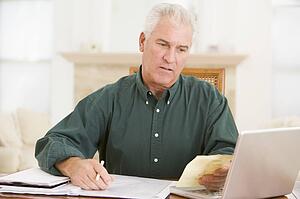 Man in dining room with laptop and paperwork looking unhappy