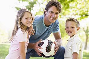 Man and two young children outdoors holding volleyball and smiling