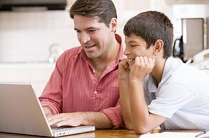 Man helping young boy in kitchen with laptop smiling