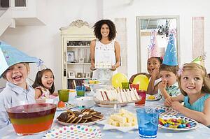 Young children at party sitting at table with mother carrying cake and smiling