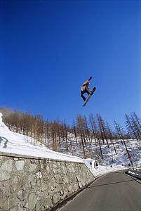 Snowboarder doing jump on snowy hill over a road (far away)