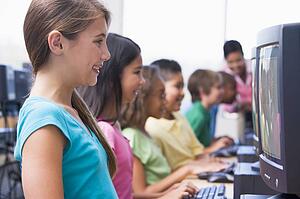 Six children at computer terminals with teacher in background (depth of field/high key)