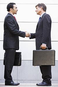 Two businessmen shaking hands outdoors holding briefcases and smiling