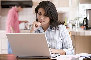 Woman in kitchen with paperwork using laptop with man in background
