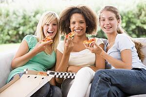 Teenage Girls Sitting On Couch And Eating Pizza Together