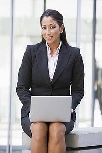 Businesswoman sitting outdoors by building with laptop smiling (high key/selective focus)