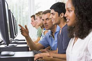 Four students sitting at computer terminals with teacher helping one of them (depth of field/high key)