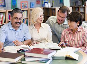 Three people sitting in library with books and notepads while a man leans over them (selective focus)