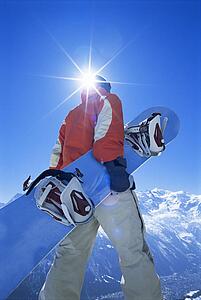 Snowboarder walking on snowy hill carrying board (lens flare)