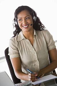 Businesswoman wearing headset in office by laptop smiling (high key/selective focus)