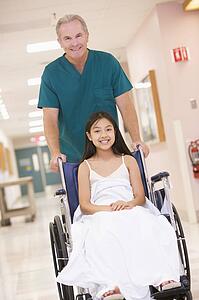 An Orderly Pushing A Little Girl In A Wheelchair Down A Hospital Corridor