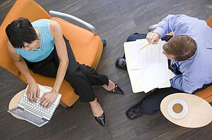 Two businesspeople sitting indoors with coffee laptop and folder