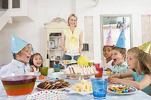 Young children at party sitting at table with mother carrying cake and smiling