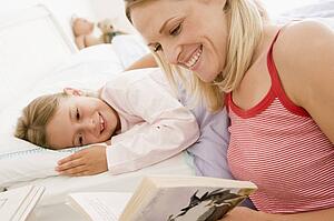 Woman reading book to young girl in bed smiling