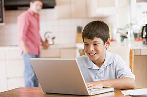 Young boy in kitchen with laptop and paperwork smiling with man in background