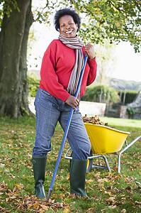 Woman outdoors raking leaves near wheelbarrow and smiling (selective focus)