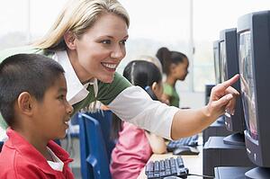 Teacher helping student at computer terminal with students in background (selective focus/high key)