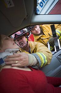 Fireman helping woman with neck brace while another fireman uses the jaws of life on a car door (selective focus)