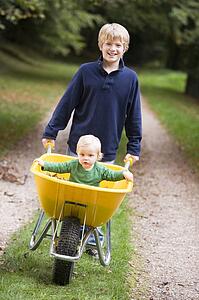 Young boy outdoors pushing baby brother in wheelbarrow and smiling (selective focus)