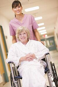 A Nurse Pushing A Senior Woman In A Wheelchair Down A Hospital Corridor