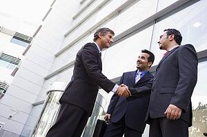 Three businessmen standing outdoors by building shaking hands and smiling (high key/selective focus)