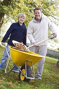 Man outdoors with young boy shoveling leaves into wheelbarrow and smiling (selective focus)