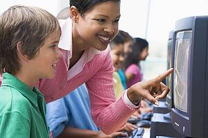 Teacher helping student at computer terminal with students in background (depth of field/high key)