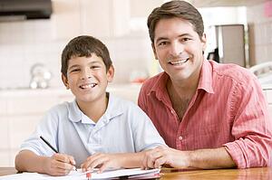 Man helping young boy in kitchen doing homework and smiling
