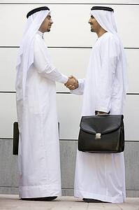 Two businessmen standing outdoors with briefcases shaking hands smiling
