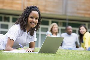 Student outdoors on lawn using laptop with other students in background (selective focus)