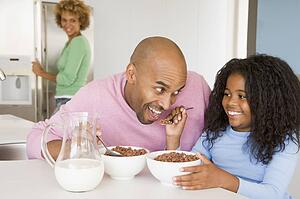 Father Sitting With Daughter As She They Eat Breakfast With Her Mother In The Background 