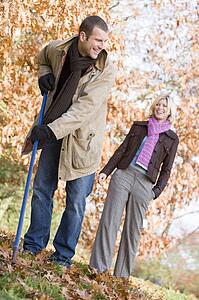Man outdoors raking leaves and woman in background smiling (selective focus)