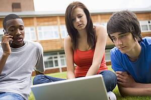 Three students outdoors on lawn with laptop one on cellular phone